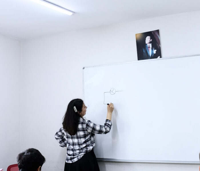 Öğretmen tahtada ders anlatıyor, öğrenciler sınıfta dinliyor